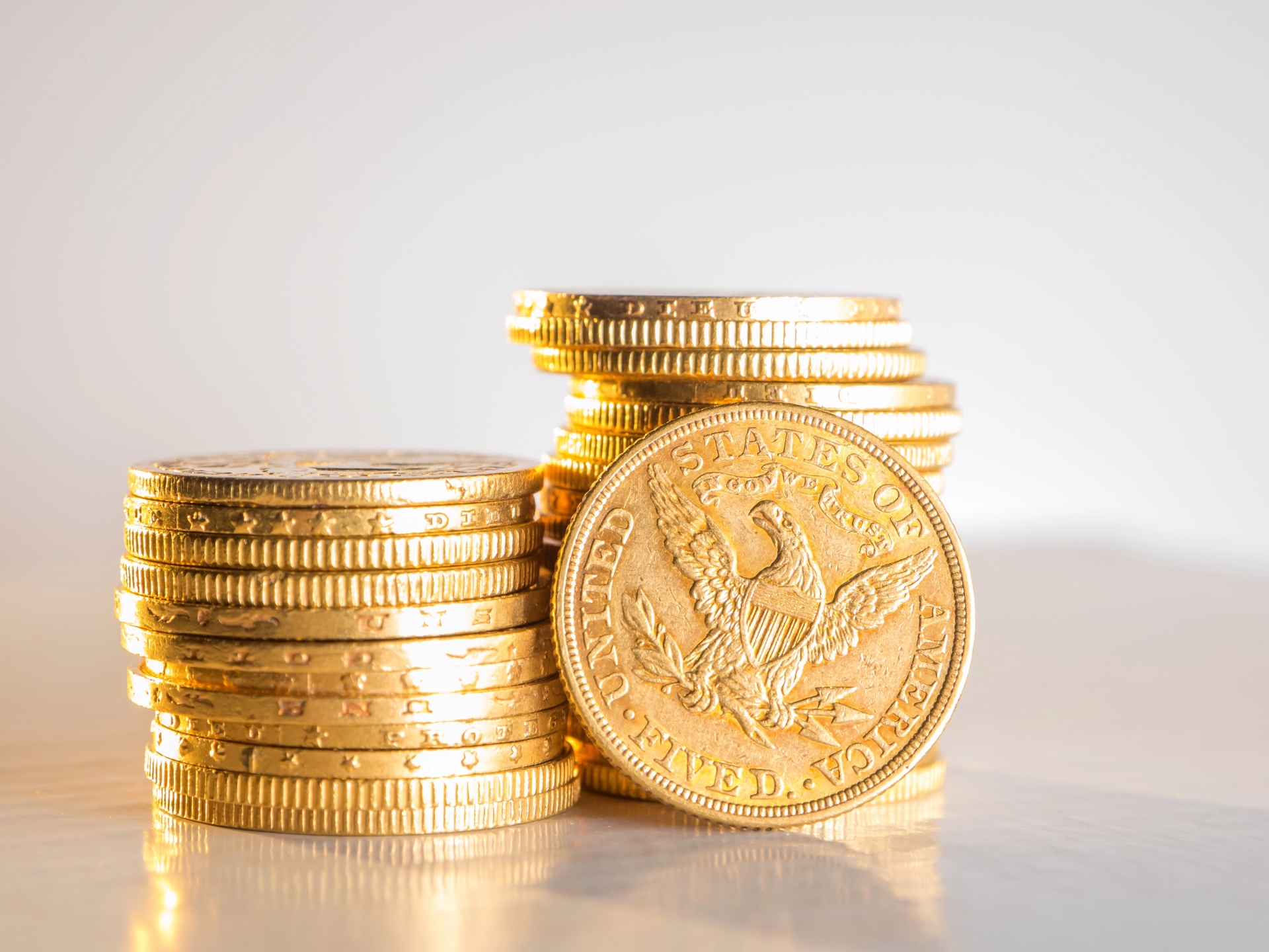 Two stacks of gold coins on a bright surface, with a five-dollar Liberty Head gold coin standing upright.