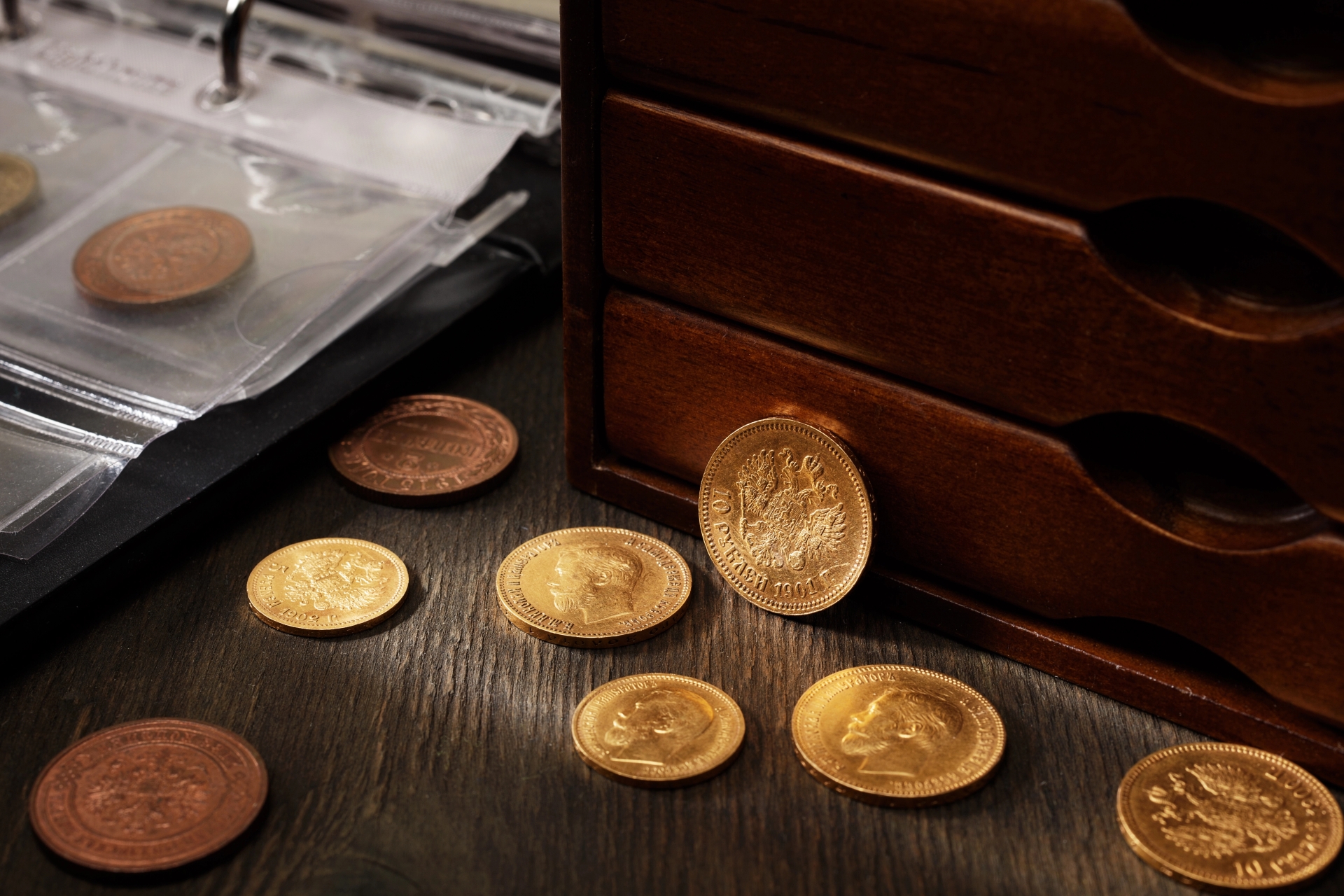 Imperial Russian gold coins scattered on a dark wooden desk next to a wooden chest and a collector's album.