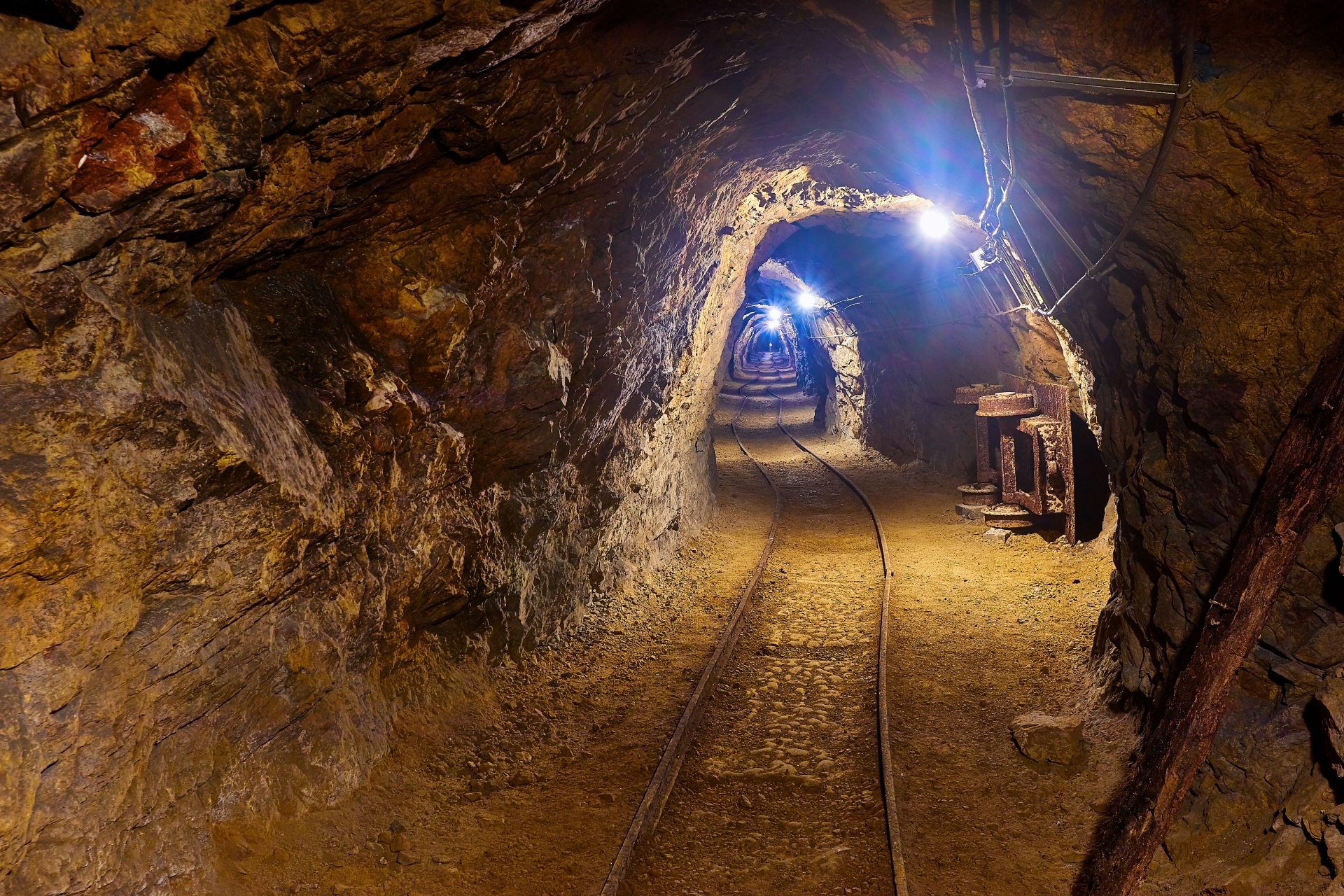 Lit underground mining tunnel showing raw rock walls and ore transport tracks used in metal extraction.