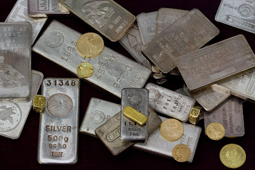 A large collection of various silver bullion bars and small gold coins scattered on a dark surface.