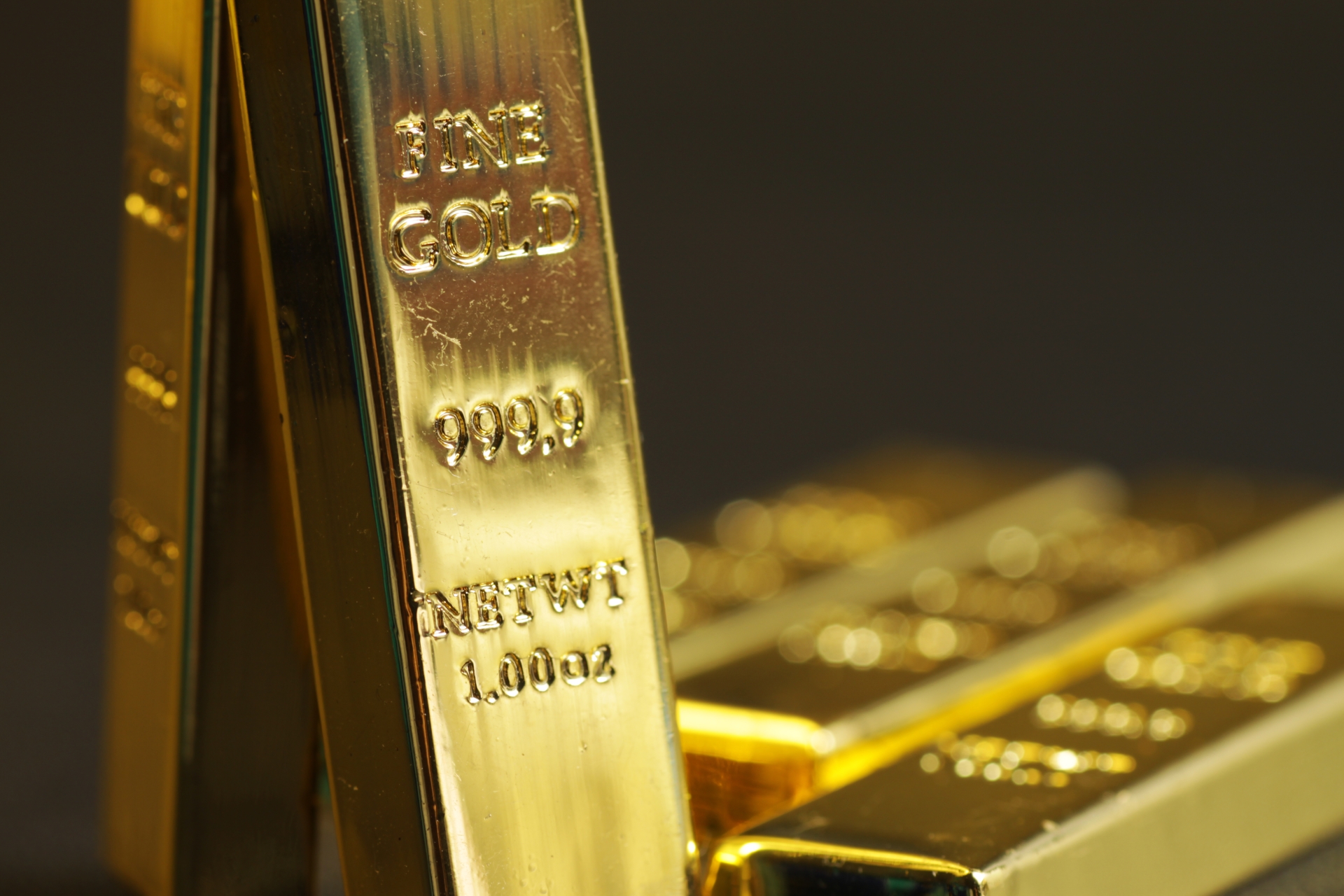 A one-ounce fine gold bar standing upright, with other gold bars blurred in the background. 
