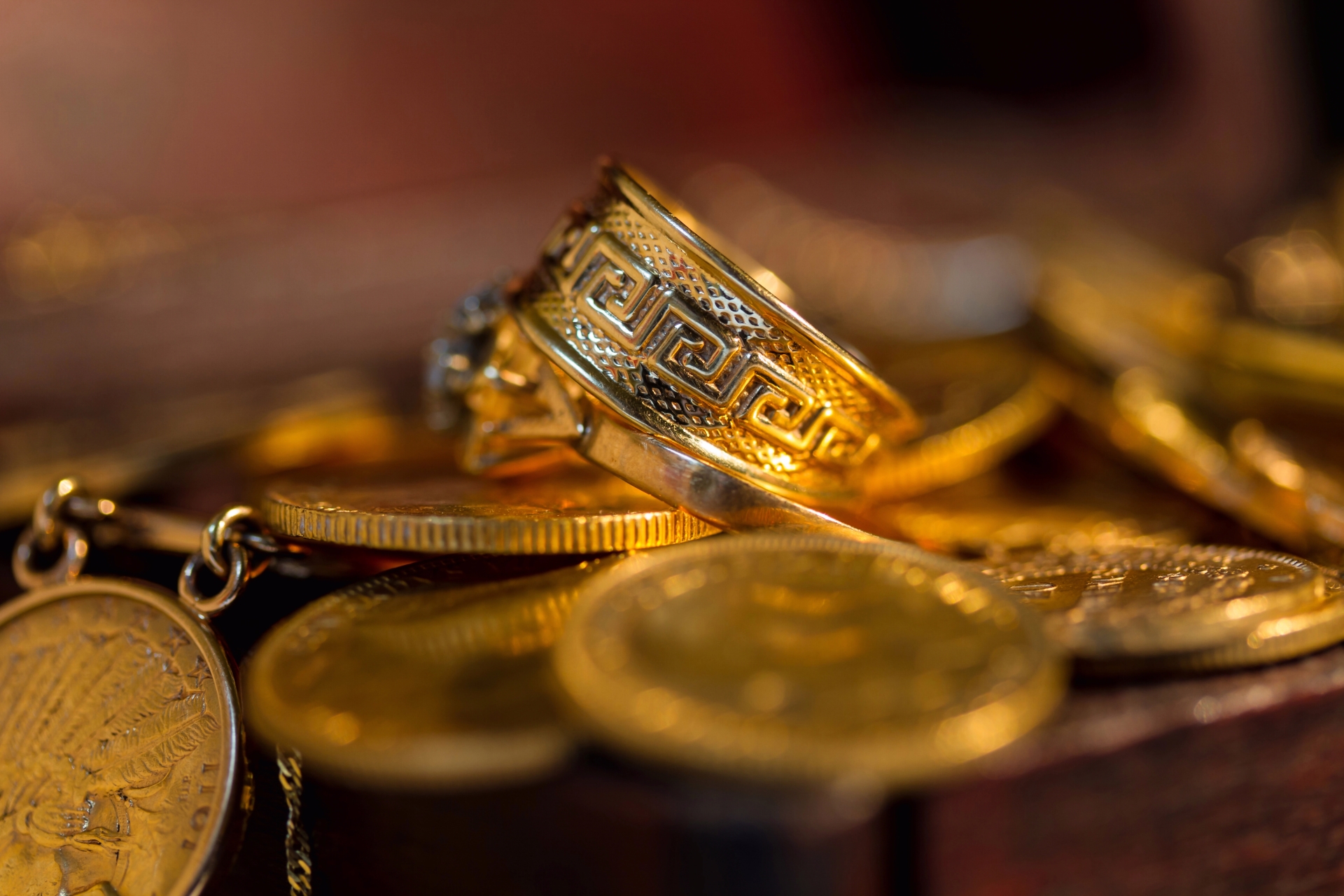 A close-up of a gold ring with a geometric design resting on a pile of gold coins and jewelry.