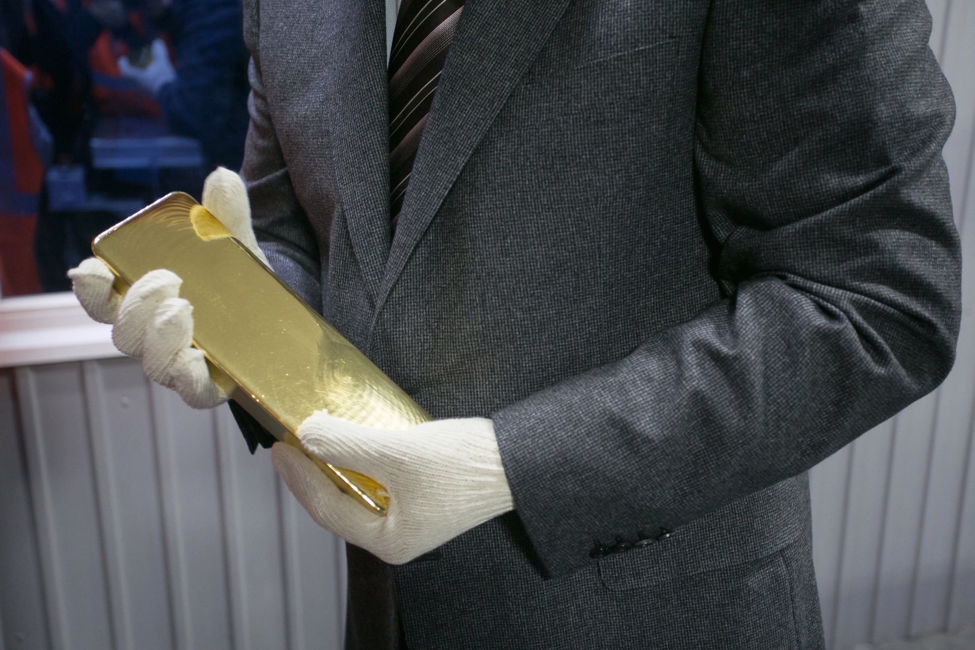 A gloved person in a suit holding a large, solid gold bullion bar in a professional setting.