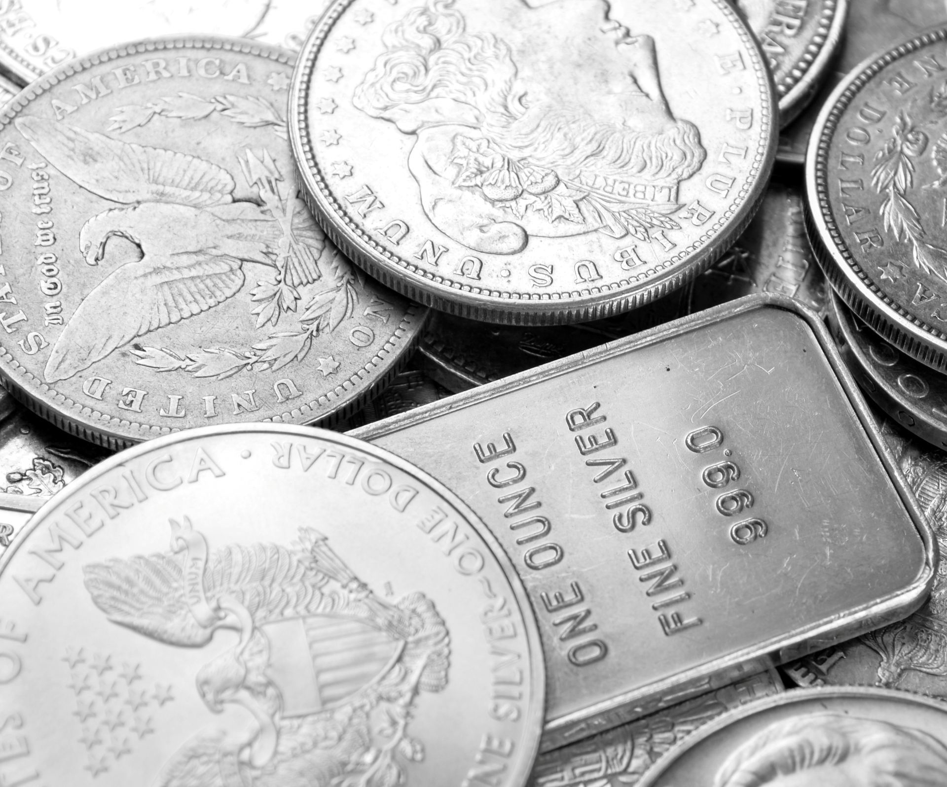 Pile of silver American Eagle and Morgan dollar coins surrounding a one-ounce .999 fine silver bullion bar.