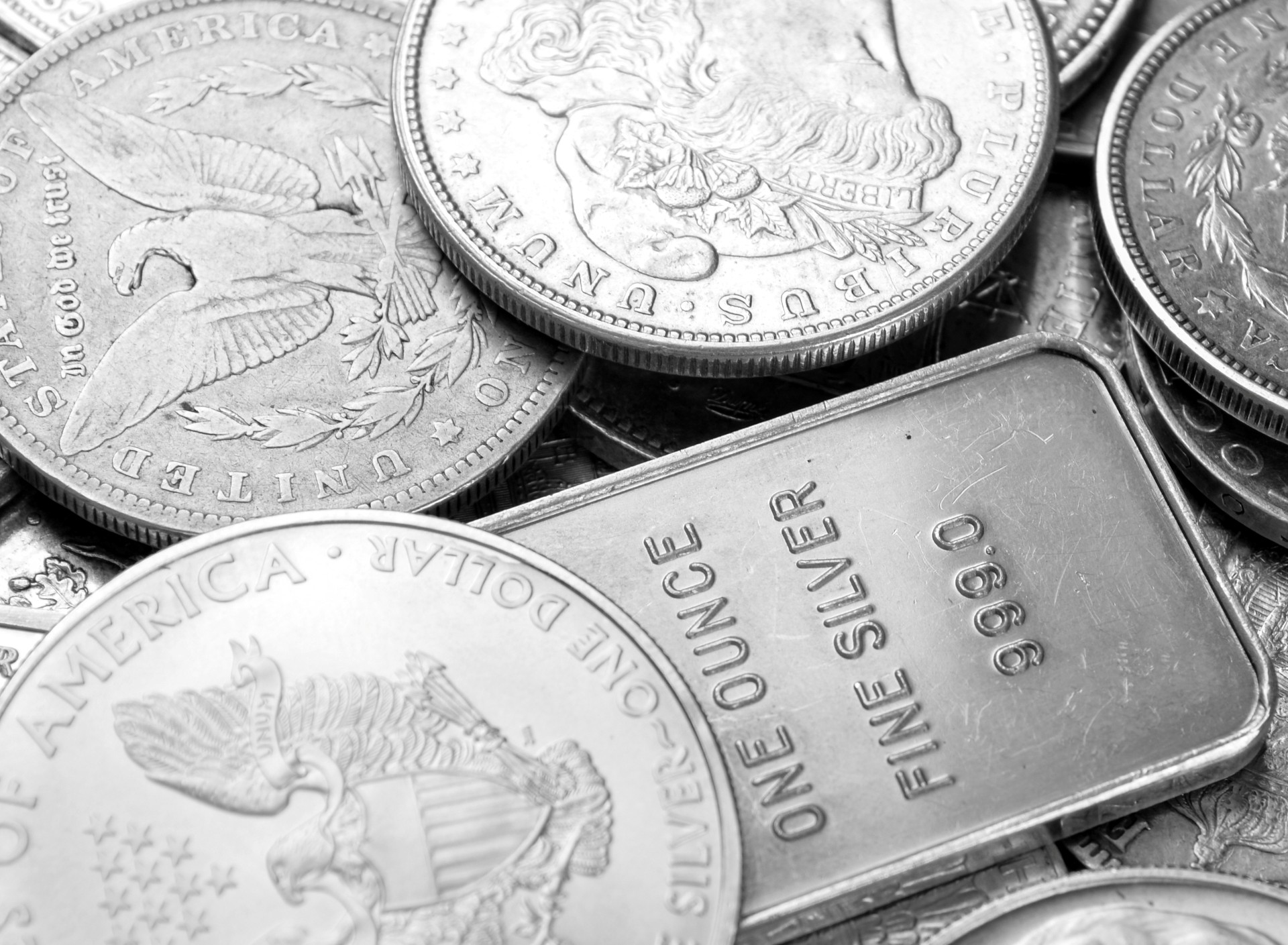 Pile of silver American Eagle and Morgan dollar coins surrounding a one-ounce .999 fine silver bullion bar.