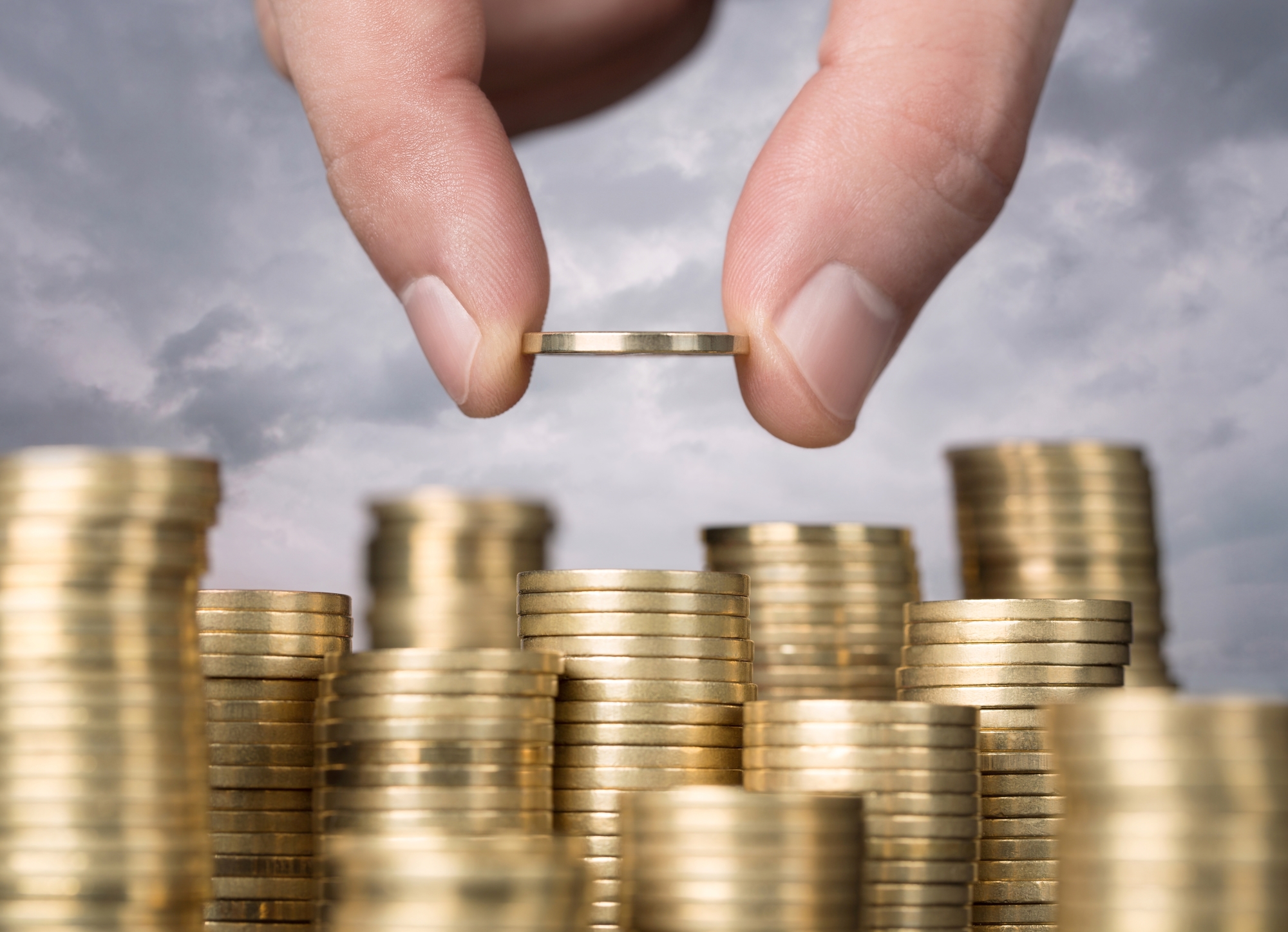 Fingers stacking gold coins with a cloudy sky background, symbolizing savings and investment growth.