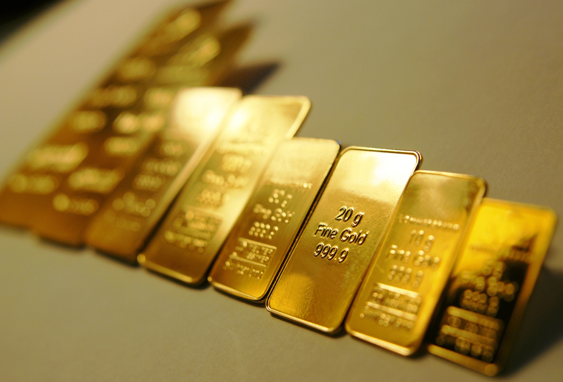 Multiple small gold bullion bars, including 10g and 20g sizes, arranged in a diagonal row on a grey surface.