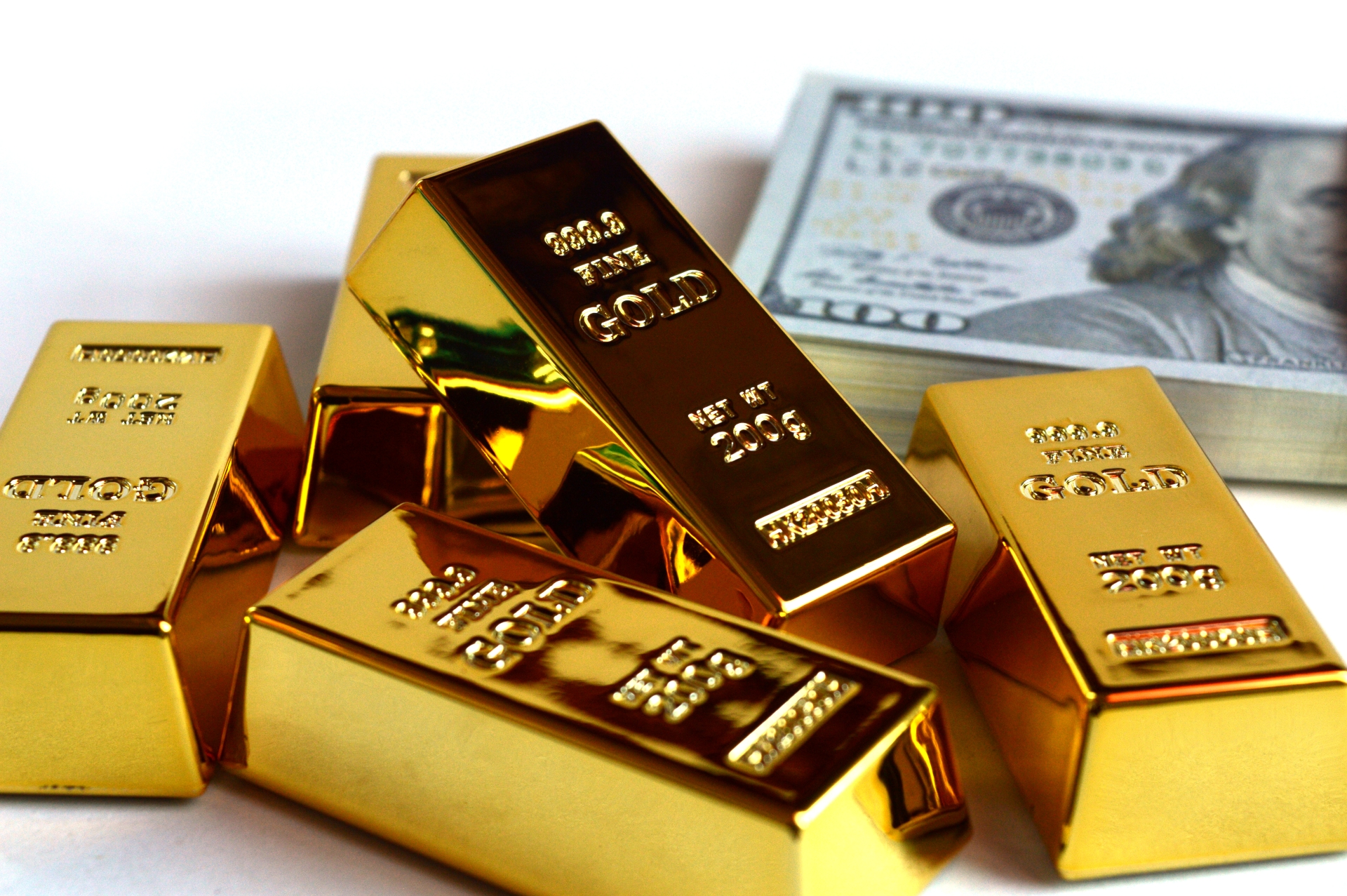 Several shiny gold bars stacked together with a bundle of U.S. hundred-dollar bills in the background.