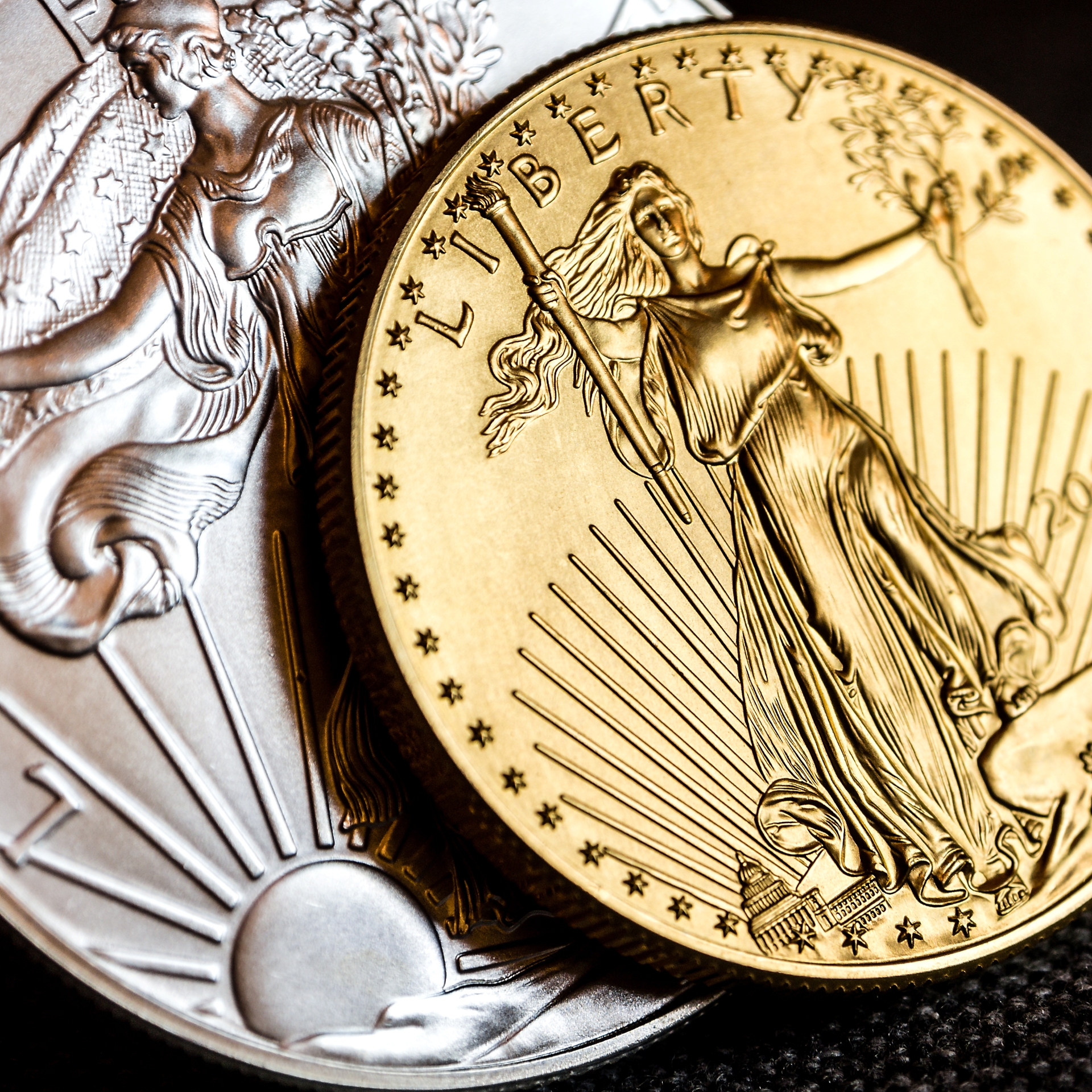 A close-up of a silver American Eagle coin next to a gold American Eagle coin, both featuring Lady Liberty.