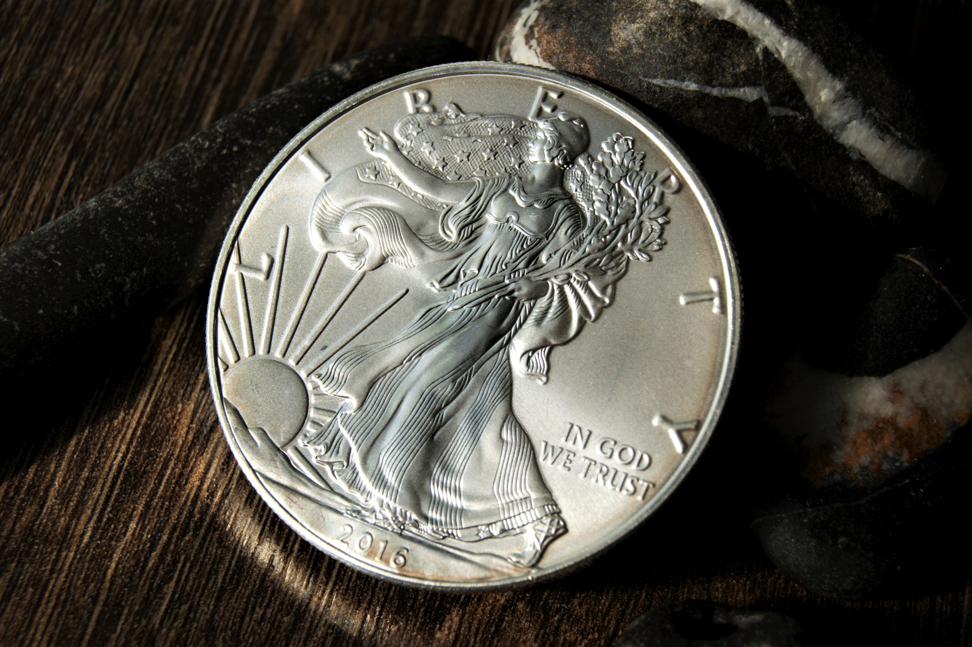 A 2016 American Silver Eagle coin resting on smooth, dark river stones and a wooden surface.