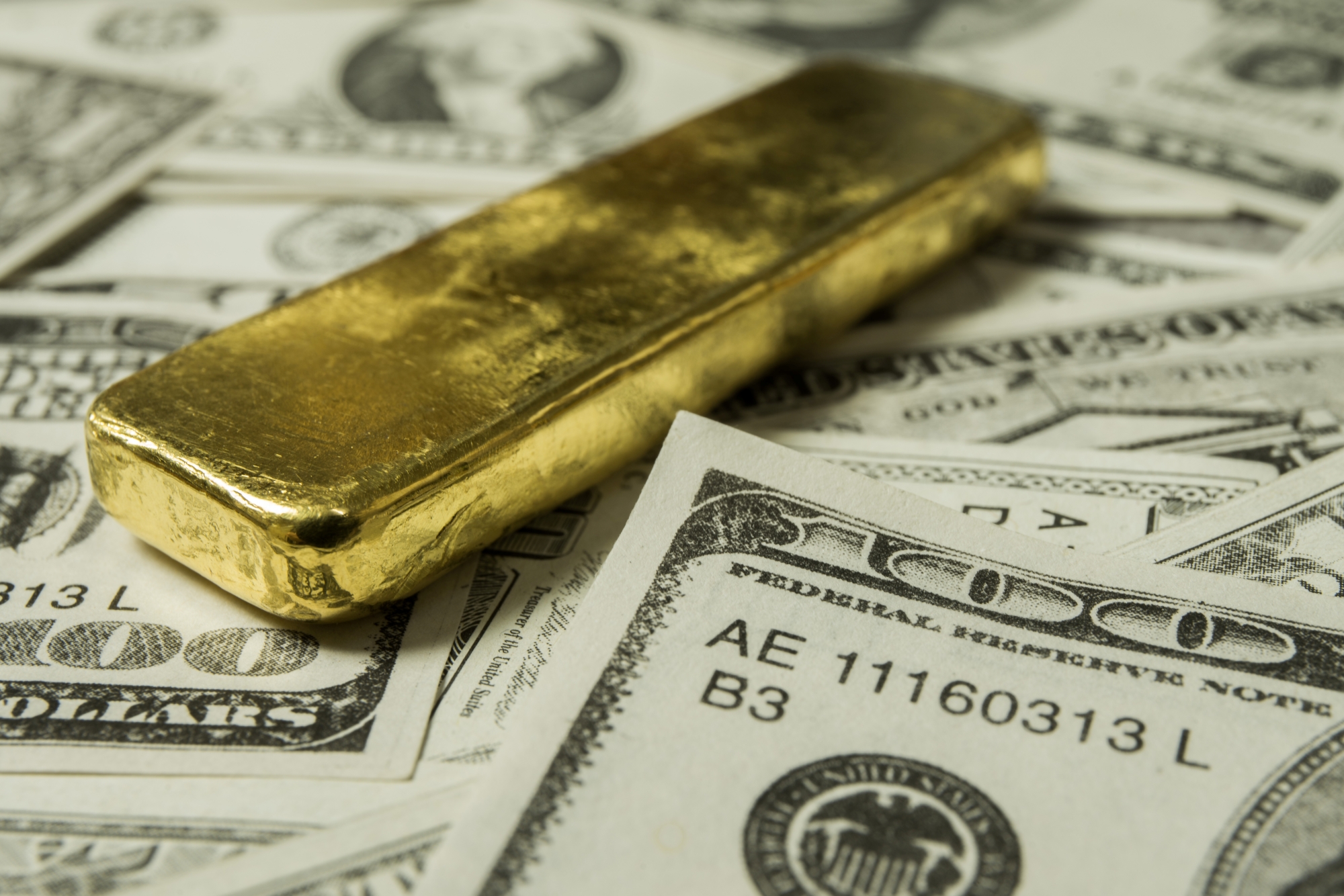 A single solid gold bullion bar resting on a scattered pile of one-hundred-dollar US federal reserve notes.