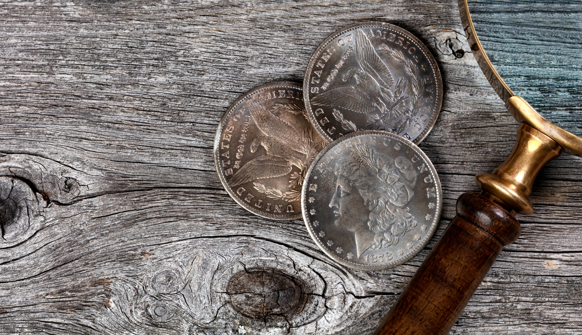 A wooden-handled magnifying glass resting next to three historic silver dollar coins on a weathered wood table.