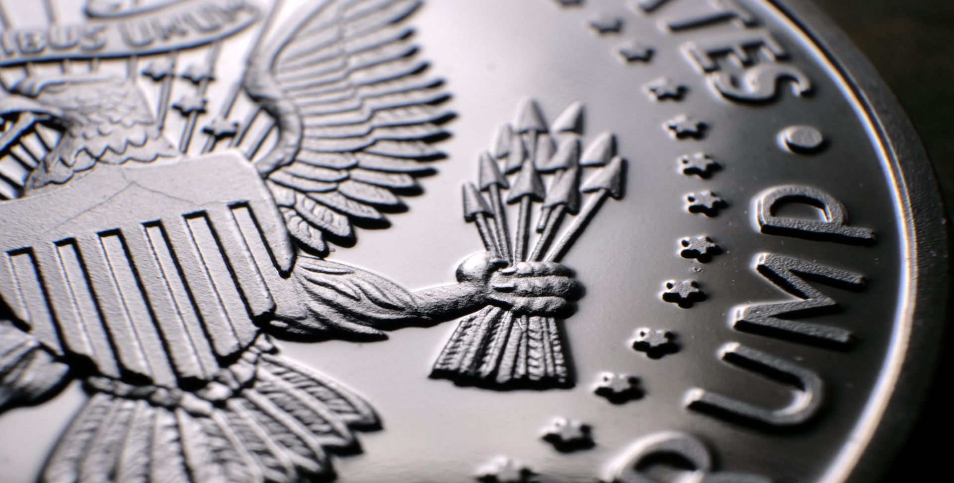 Macro close-up of a silver coin featuring a detailed eagle's wing and a hand clutching a bundle of arrows.
