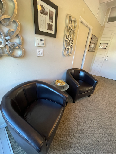 Two black leather chairs and a small glass table with decorative gold items, in a light-filled modern office with framed art on the wall.