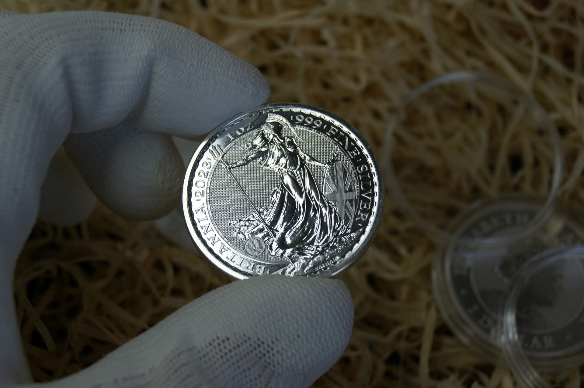 A gloved hand holding a silver Britannia coin over a bed of wood shavings and plastic coin capsules.