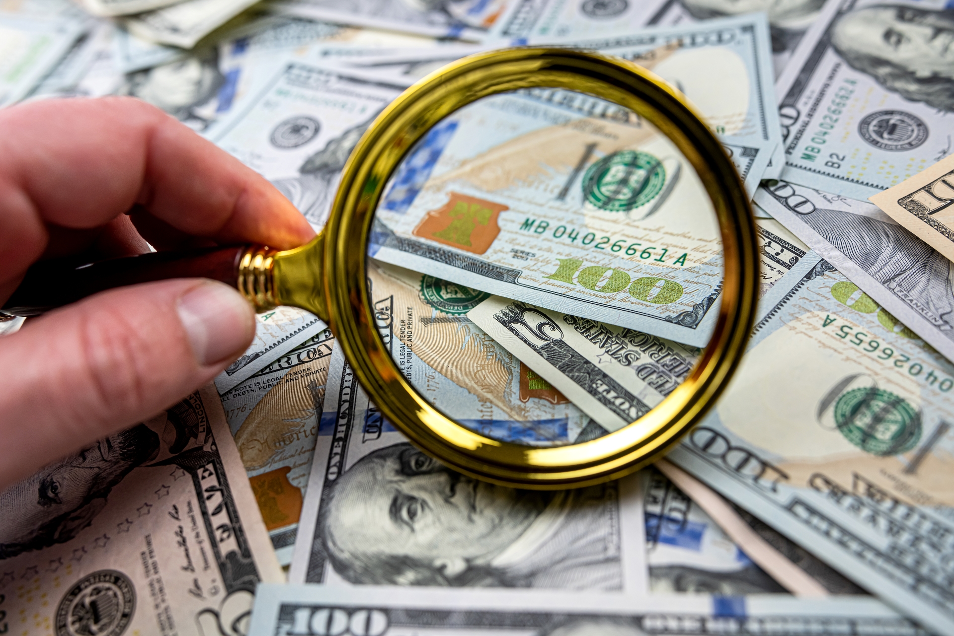 A hand holds a magnifying glass over a scattered pile of US$100 bills.